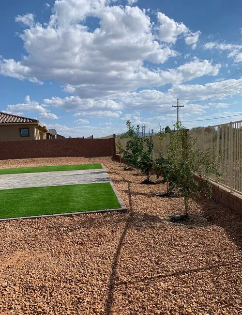 A renovated backyard enhanced by professional landscaping services, featuring a row of trees set within a spacious gravel area that surrounds a lush grass turf.
