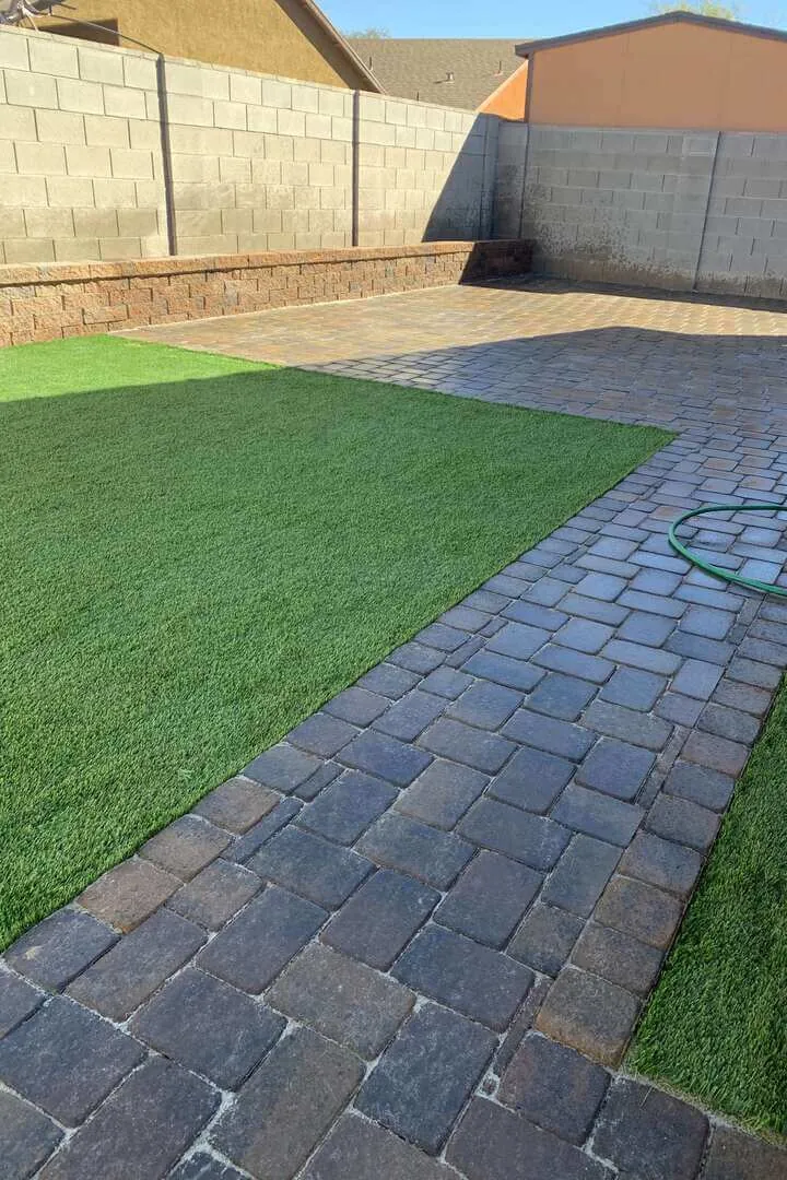 A corner shot of a backyard with a tiled patio covering most of the backyard and a small grass field in the corner, all surrounded by concrete retaining walls. The backyard had yard maintenance as it is very clean.