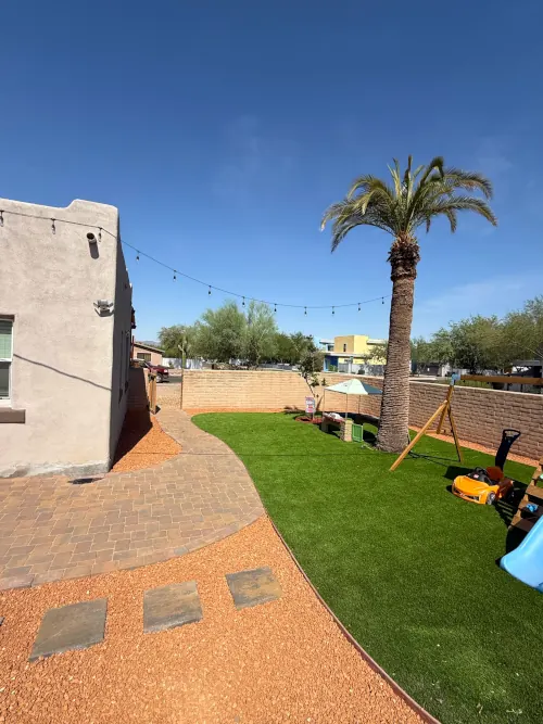 Newly installed artificial grass lawn with curved paver walkway, decomposed granite path with stepping stones, and a palm tree in a sunny desert backyard.
